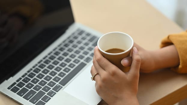 Cropped shot young woman holding coffee cup and using laptop on wooden desk.