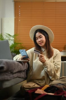 Happy young woman holding credit card and using mobile phone booking tickets or hotel room on web.