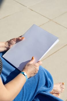 reading at vacation. unrecognizable woman holding blank magazine for mockup design, sitting by the swimming pool in blue towel