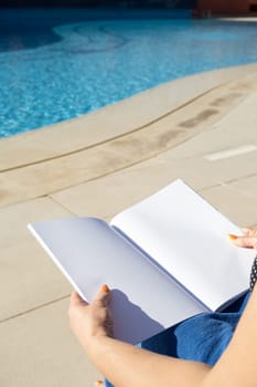 reading at vacation. unrecognizable woman holding blank magazine for mockup design, sitting by the swimming pool in blue towel