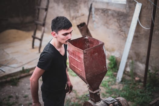 One young Caucasian brunette guy in an old and dirty black T-shirt looks into a homemade crusher where dry animal feed is crushed, standing near a village house, top side close-up view in dark style.