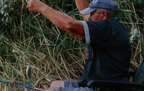 One Caucasian elderly man in old dirty clothes pulls out a fish caught with a fishing rod from the river while sitting on a chair on the shore of a lake on a sunny summer day, close-up side view.