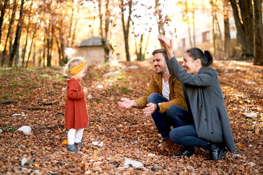 Smiling mom and dad sprinkle dry leaves on a little girl sitting on the ground in the autumn forest. High quality photo