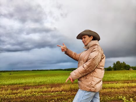 An adult girl looking like a cowboy in a hat in a field and with a stormy sky with clouds takes pictures of rainbow and takes selfie in the rain. Woman having fun outdoors on rural and rustic nature