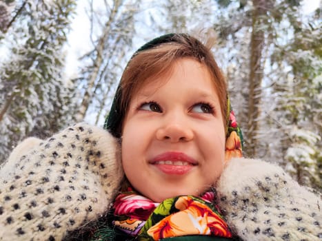 Cute girl in a traditional Russian headscarf and mittens on winter forest. Closeup portrait of child in folk clothes. Carnival Maslenitsa