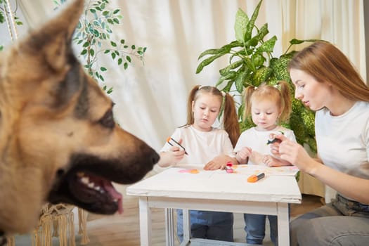 Young mother or babysitter, little daughter, sister teenager girl drawing at table and big dog shepherd. Family enjoying leisure at home