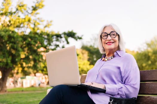 smiling senior woman using laptop outdoors sitting on park bench, concept of technology and elderly people leisure, copy space for text