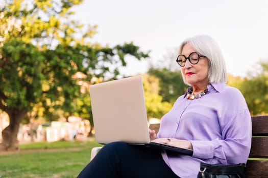 senior woman using laptop outdoors sitting on park bench, concept of technology and elderly people leisure, copy space for text