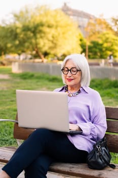 beautiful senior woman with glasses using laptop outdoors sitting on park bench, concept of technology and elderly people leisure, copy space for text