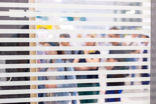 Blurred figures of people seen through the slats of window blinds, creating an abstract effect.