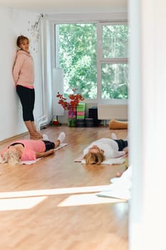 A skilled trainer oversees a group of senior women practicing various yoga exercises, including neck, back, and leg stretches, in a sunlit space, promoting wellness and harmony.