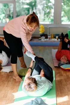 A skilled trainer oversees a group of senior women practicing various yoga exercises, including neck, back, and leg stretches, in a sunlit space, promoting wellness and harmony.