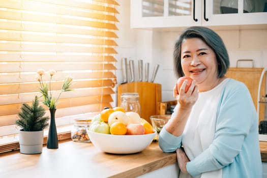 A smiling old Asian woman, a grandmother, finds joy in the kitchen, cooking and preparing healthy vegan food. She selects a fresh apple for eating, emphasizing the diet concept and happiness.