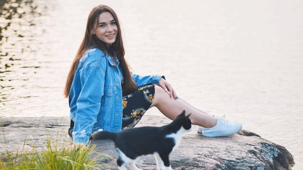 A girl sits on a rock with a cat against the background of a lake