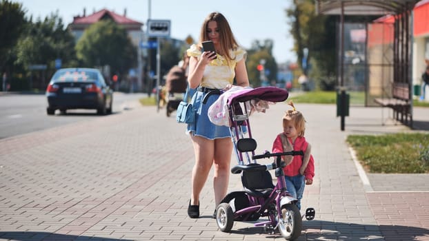 A young mother with a baby and a pram walks through town and looks at her phone