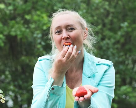 Portrait of a beautiful Caucasian cheerful middle-aged blonde woman eating large ripe strawberries with appetizing pleasure while sitting in a park on a bench on a spring day, close-up side view. Outdoor picnic concept, healthy eating and lifestyle.