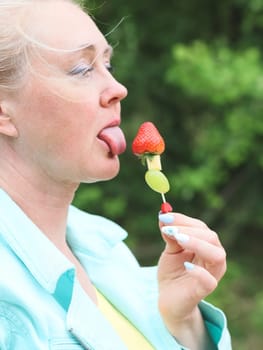 One Caucasian young beautiful blonde woman with delicate manicure in a blue jacket and yellow T-shirt holds licking one fruit barbecue skewer of strawberries, cheese and wongrad in the park on a spring day, side view close-up. Spring picnic concept, healthy eating.