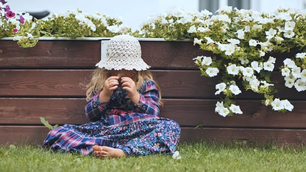 A little girl in a panama hat sits by the white petunias in the garden