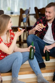 The couple is having dinner at home. They hold slices of pizza. The man is sitting forward and can be seen smiling. They are both holding green bottles of beer.