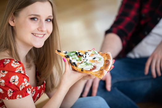 Top view. A young girl holds a slice of pizza in front of her. She smiles serenely. Blurred background. A fragment of a male silhouette can be seen in the background.