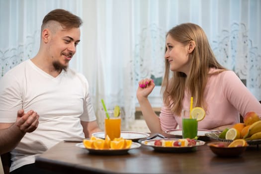 A man in a white t-shirt is talking about something, gesturing. A young girl in a pink sweatshirt sits next to him and listens to him. The couple sits at a table with lots of fruit on it.