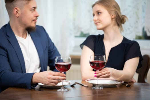 A man and a woman sit at a table. They are waiting for a meal. In front of them on the table lie two empty plates. The couple is holding glasses of red wine.