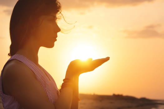 Portrait of a young woman in the form of a silhouette and hands holding the sun. Sunset over the sea, the sun on the palms of a calm girl.