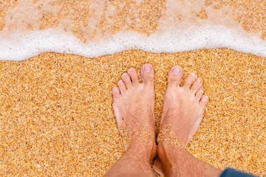 Top view of male legs in blue shorts on golden sand in sunlight. Looking down at a man in shorts standing on the sea sandy beach.