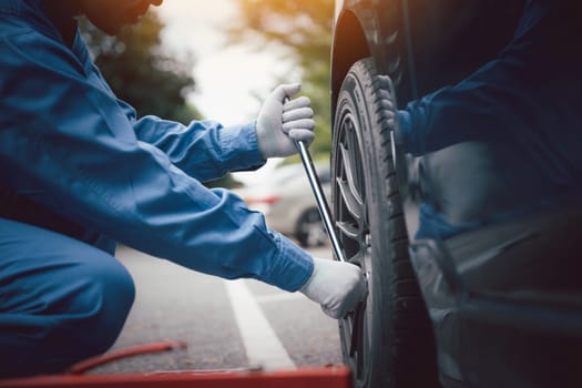 Skilled car mechanic in a blue work uniform, squatting next to a vehicle in an auto repair shop, fixing a tire with a focus on the hands.