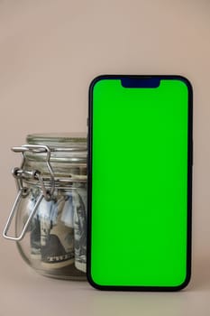 Chrome key smartphone screen mock up template in vertical position on beige background. Copy space App website advertising. Jar filled with dollars cash. Concept of Mobile application and technology business savings.