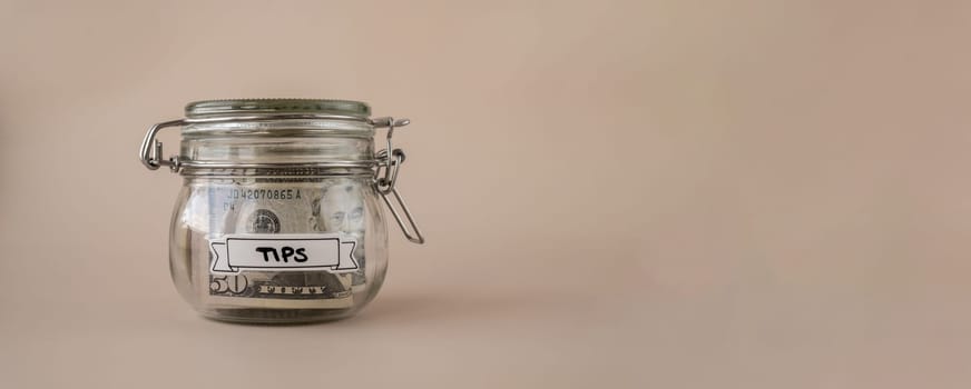 Saving Money In Glass Jar filled with Dollars banknotes. TIPS transcription in front of jar. Managing personal finances extra income for future insecurity. Beige background