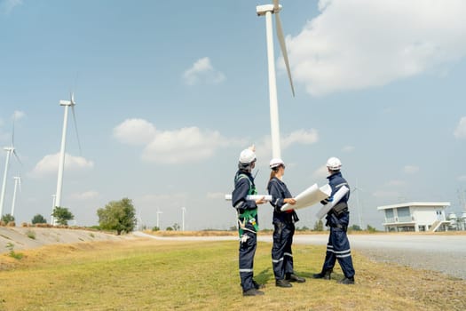 Group of professional technician workers stand with holding project paper plan in front of wind turbine or windmill and look back in area for power plant business.