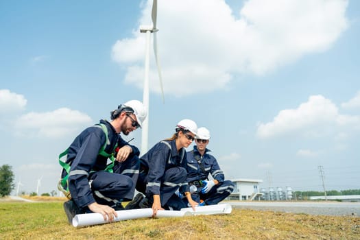 Side view of group professional technician workers sit and discuss work with project paper plan in front of wind turbine or windmill and point to the pole in area for power plant business.