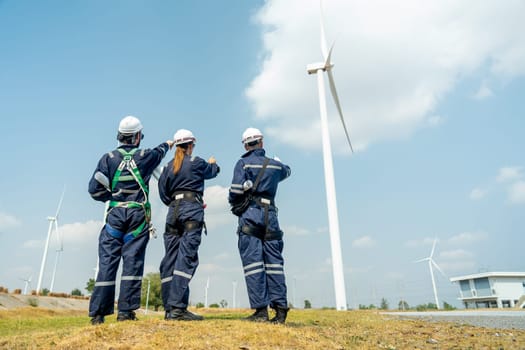 Back of group of professional technician man and woman workers with safety suit stand in front of wind turbine or windmill and point to the pole in area for power plant business.