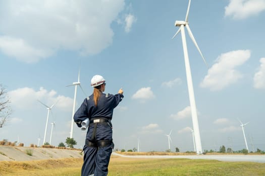 Professional technician woman worker stand in front of wind turbine or windmill and point to the pole in area for power plant business.