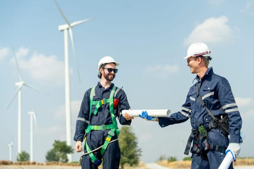 Professional Caucasian workers or technician with safety suit hold and send the construction paper plan to co-worker also stand in front of wind turbine or windmill in the row as power plant business.