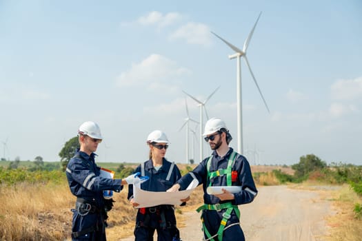 Group of professional technician man and woman workers discuss about work project on plan paper in front of wind turbine or windmill in workplace area.