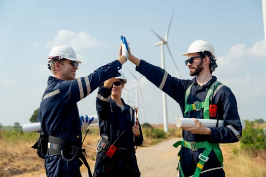 Close up professional technician workers men and woman stand with touch hands in front of roll of wind turbine or windmill in the field area of power plant business.