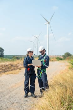 Two technician workers man and woman discuss about work using laptop and stand in front of roll of wind turbine or windmill in the field area of power plant business.