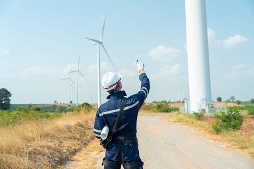 Back of technician worker man stand and point up to the sky and wind turbine or windmill on the back in area of power plant business.