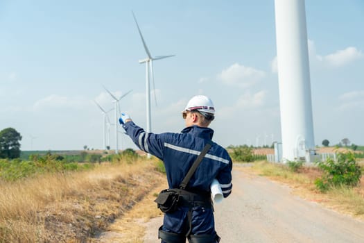 Back of technician worker man stand and point to wind turbine or windmill on the back in area of power plant business.