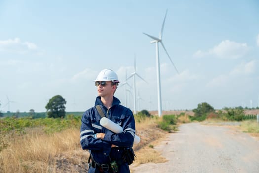 Professional technician worker man stand with arm-crossed and stand with look to his right side in front of wind turbine or windmill in the field and in concept of green energy for good environment.