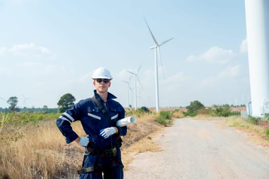 Professional technician worker man stand with hold plan paper and look forward in front of wind turbine or windmill with blue sky in concept of green energy for good environment.