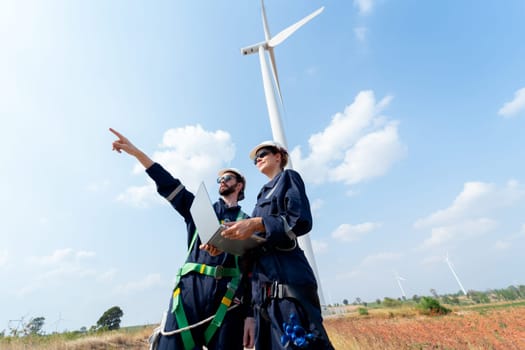 Professional worker woman hold laptop and stand with her co-worker man point forward and they stand in front of wind turbine or windmill in the field for power plant business.