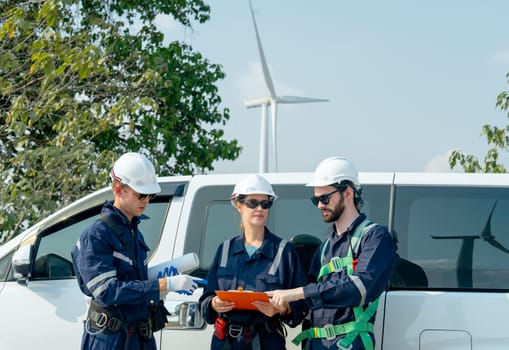 Group of technician workers stand beside the van and discuss about work with windmill or wind turbine on the background.