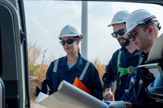 Close up Group of technician workers stand beside the van that open the door and discuss about work using plan paper with windmill or wind turbine on the background.