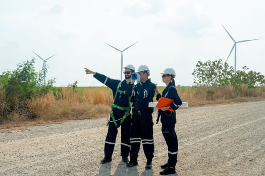Group of technician workers stand on the road and discuss about work one of them also point to left side with windmill or wind turbine on the background.