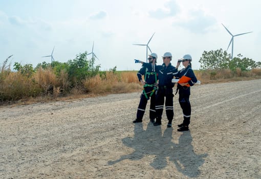 Wide short group of technician workers stand on the road and discuss about work with one of them point to left side and stay with windmill or wind turbine on the background.