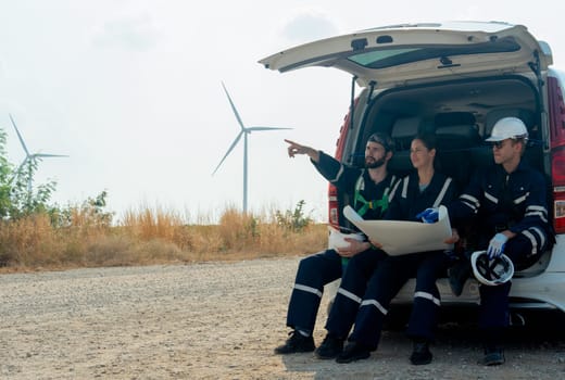 Side view group of technician workers sit on back of van and discuss about work in plan paper on the road with windmill or wind turbine on the background.