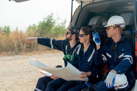 Group of technician workers sit on back of van and discuss about work in plan paper and one point to left side stay on the road in area of windmill or wind turbine on the background.